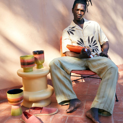 Person sitting on a chair with colorful stools and cups on a red brick floor.