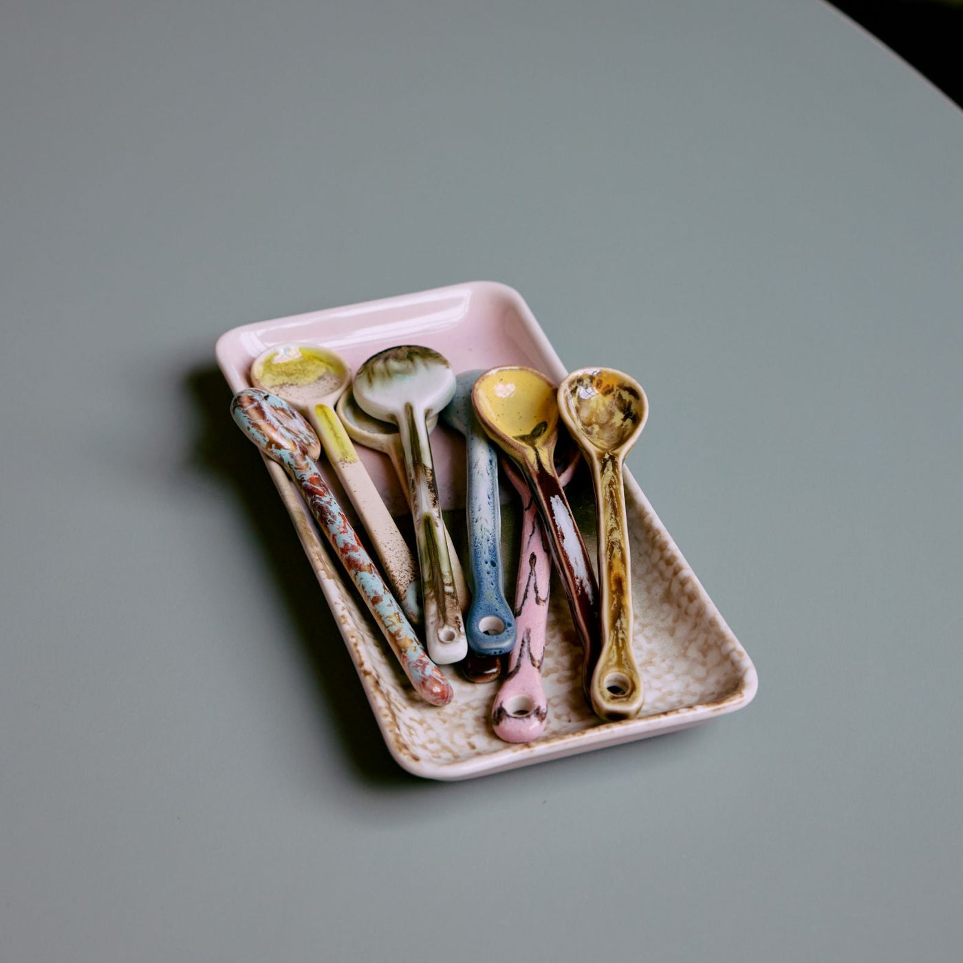 Set of colorful ceramic spoons on a pink tray on a gray surface.