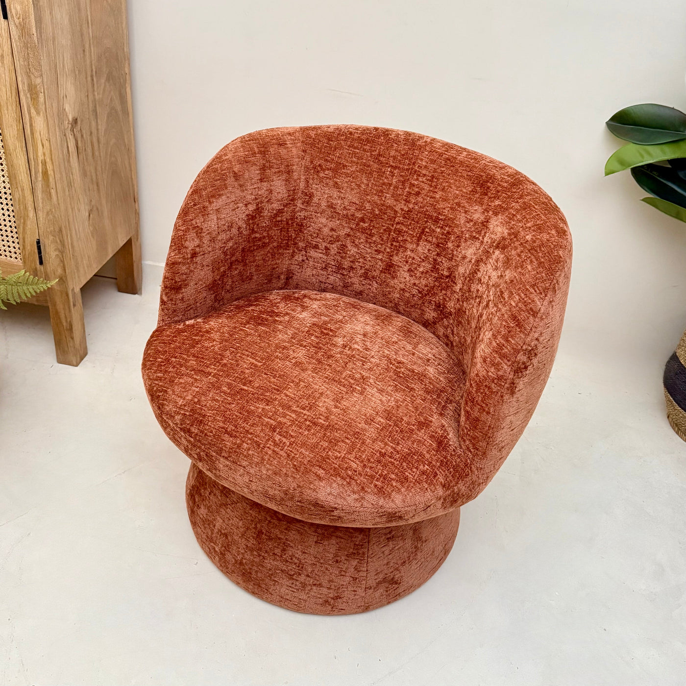 Terracotta chenille swivel armchair in a room with a wooden cabinet and plants.-Mrs Robinson