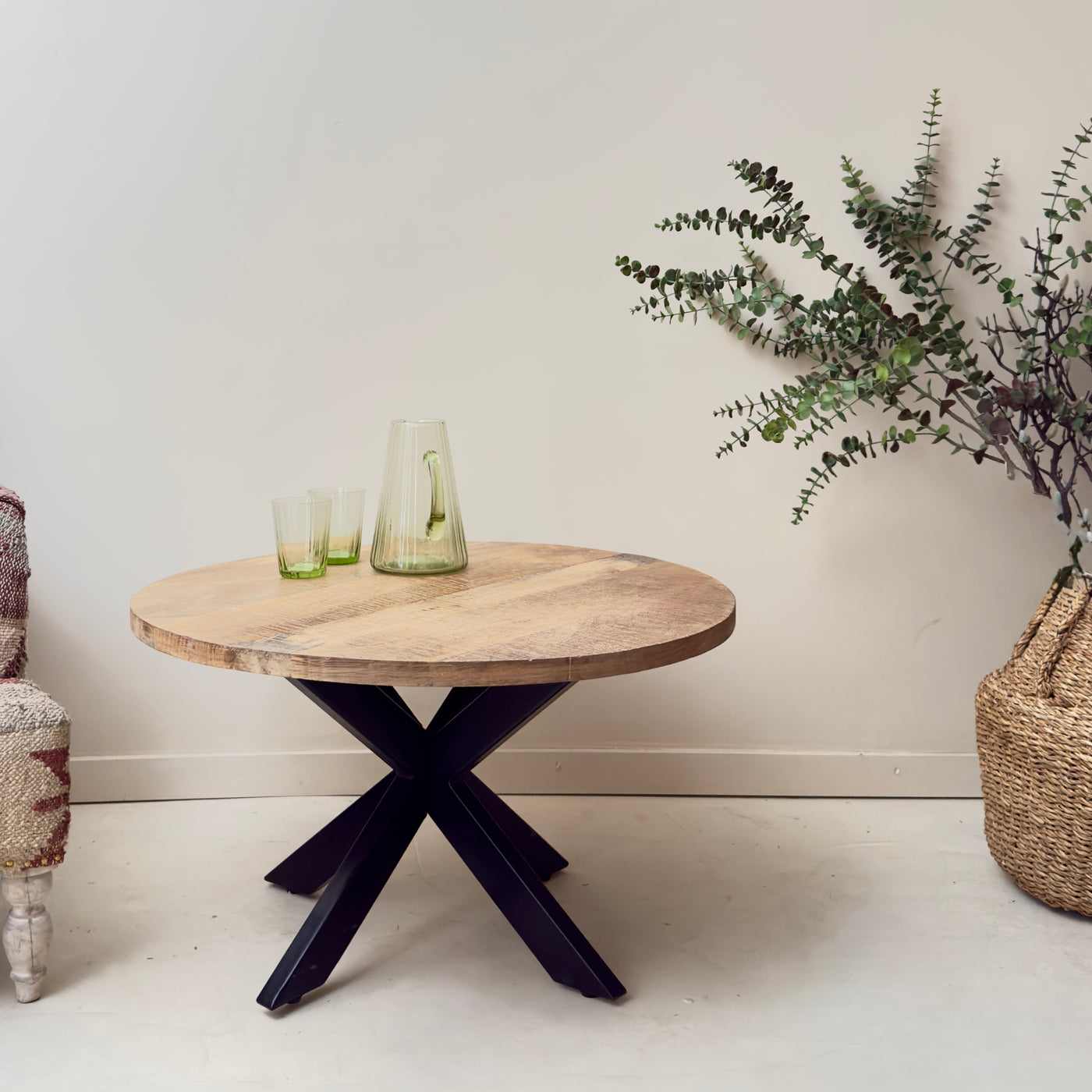 Round wooden table with black metal legs in a minimalistic room with a plant and vase.