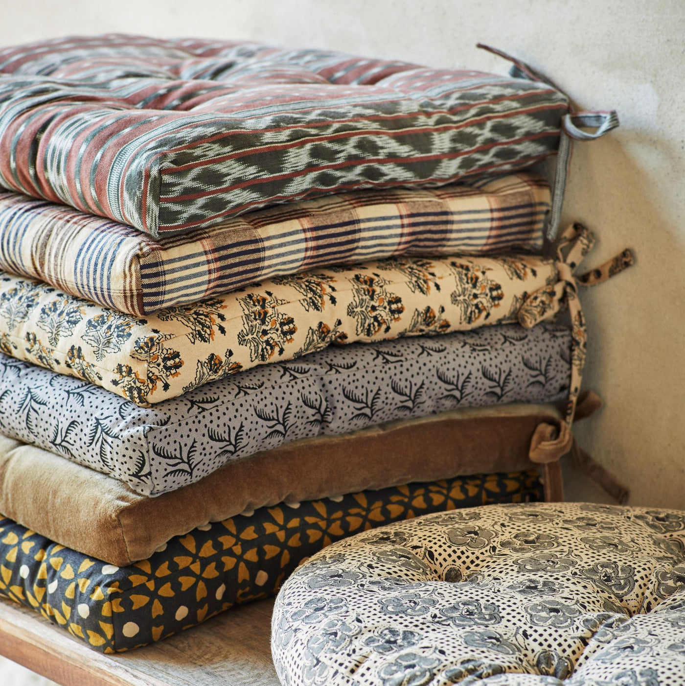 Stack of patterned cushions on a wooden bench against a neutral wall.